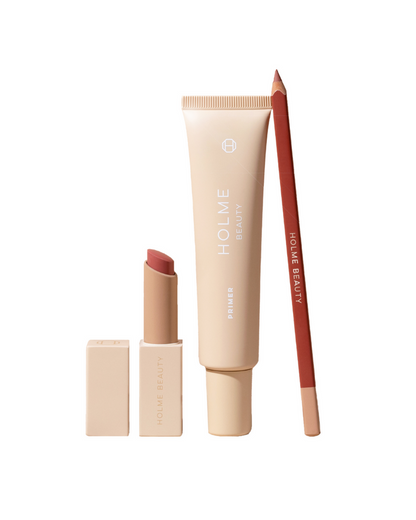 A collection of three makeup products including a tube labeled 'HOLME BEAUTY' with a cap, a stick of lip liner, and a tube of lipstick, all in a neutral skin tone color range.